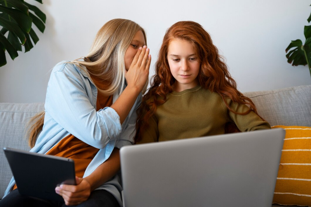 Blonde young lady whispering to a redheaded young lady looking at a computer screen