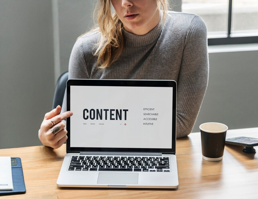 Blonde lady in a grey sweater pointing at a computer screen with a website up labeled "Content" lots of white space, and sublinks clearly defined. 